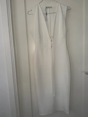 Vit långklänning med snörning från Aéryne - Elegant vit långklänning från Aéryne med djup v-ringning och snygg snörningsdetalj framtill. Klänningen är ärmlös och har en stilren, figurnära passform. Perfekt för dig som vill ha en minimalistisk och modern look.