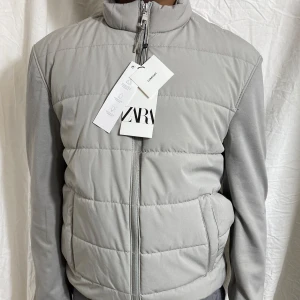 Ljusgrå quiltad väst från ZARA - Säljer en ljusgrå quiltad väst från ZARA med dragkedja framtill och ståkrage. Västen har två sidofickor och en stilren, minimalistisk design. Perfekt för lager-på-lager och enkel att matcha med olika outfits.