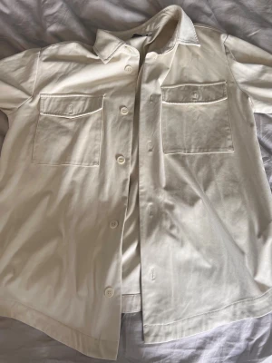 Kritvit skjorta med fickor - Stilren kritvit skjorta med två stora bröstfickor och klassisk krage. Skjortan har raka ärmar, knäppning framtill och är gjord i ett mjukt, lätt material som känns skönt mot huden. Perfekt för en clean och enkel look.