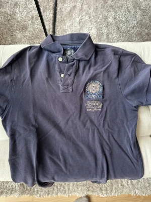 Marinblå pikétröja från Morris - Klassisk marinblå pikétröja från Morris med broderad logga och emblem på bröstet. Tröjan har krage, knappar framtill och korta ärmar. Perfekt för en stilren och sportig look.