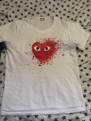 Vit Comme des Garçons Play t-shirt - Vit t-shirt från Comme des Garçons Play med det ikoniska röda hjärtat med ögon på bröstet och stänkmönster runt motivet. Klassisk rund hals och korta ärmar. Perfekt för dig som gillar streetwear och vill sticka ut.