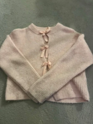 Ljusrosa kofta med rosetter - Supersöt ljusrosa kofta i ribbstickat material med tre dekorativa rosetter framtill. Koftan har rund halsringning och långa ärmar, perfekt för en gullig och trendig look.