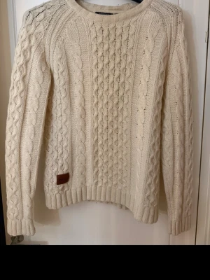 Krämvit kabelstickad tröja WEZC - Snygg kabelstickad tröja från WEZC i krämvit färg. Tröjan har rund halsringning, ribbade muddar och ett klassiskt flätmönster. Perfekt för lager-på-lager och chill dagar. Liten läderpatch med logga nertill.