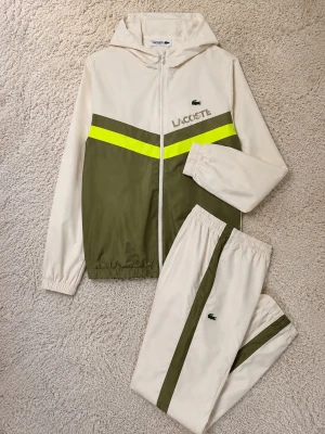 Lacoste tracksuit - Tracksuit från Lacoste i beige och olivgrönt med en neon-gul rand över bröstet. Jackan har huva, dragkedja och elastiska muddar. Byxorna har matchande olivgrön och neon-gul detalj längs benet. Perfekt för sport eller chill.