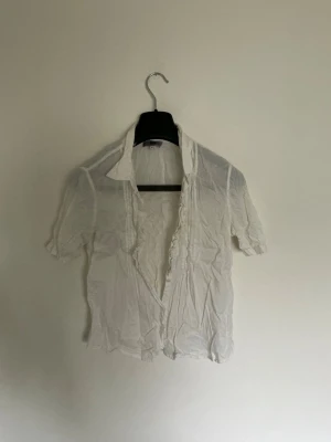 Vit blus med volangkant - Snygg vit blus med korta ärmar och volangkant längs knäppningen. Blusen har klassisk krage och är tillverkad i ett tunt, lätt material som ger en luftig känsla. Perfekt att styla med jeans eller kjol för en fräsch look.