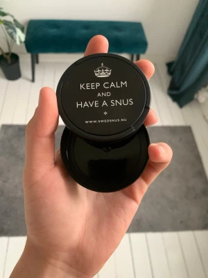 Svart snusdosa med text - Säljer en svart snusdosa i plast från Swedsnus med texten 'KEEP CALM AND HAVE A SNUS' på locket. Dosan är rund och har ett enkelt, stilrent utseende. Perfekt för att förvara snus på ett smidigt sätt.