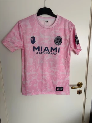 Rosa BAPE x Inter Miami fotbollströja - Säljer en limited edition rosa fotbollströja från BAPE x Inter Miami med kamouflagemönster, tryckta loggor och texten MIAMI på bröstet. Nummer 93 och BAPE på ryggen. Korta ärmar och rund hals. Perfekt för dig som gillar streetwear och fotboll.