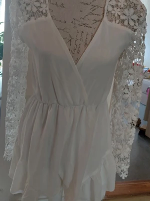 Vit jumpsuit med spetsärmar - Supersöt vit jumpsuit med omlottliv och långa ärmar i blommig spets. Resår i midjan och volangdetaljer nertill. Perfekt för dig som vill ha en romantisk och trendig look. Materialet känns lätt och luftigt.