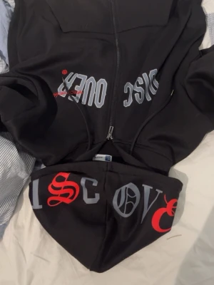 Svart hoodie med tryck och dragkedja - Svart hoodie med dragkedja och stor huva. Tryck i grått och rött på både bröstet och huvan, där det står 'DISCOVER' i gotisk stil. Hoodien har en avslappnad passform och är tillverkad i mjukt bomullsmaterial.