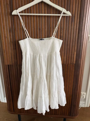 Vit sommarklänning med smock - Luftig vit klänning från Zara med smock upptill och tunna axelband. Klänningen har en volangig kjol och är tillverkad i ett lätt bomullstyg, perfekt för varma dagar. 