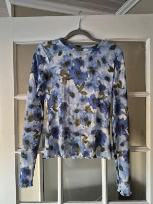 Blommig ribbad långärmad topp - Superfin långärmad topp med blått och grönt blommönster på vit botten. Ribbad struktur och lätt croppad passform. Perfekt att styla med jeans eller kjol för en trendig look.