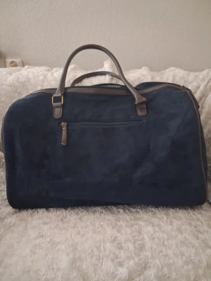 Mörkblå weekendbag i mocka-look - Snygg mörkblå Hunton weekendbag med mocka-liknande yta och bruna handtag i läderimitation. Väskan har en ytterficka med dragkedja och ett mönstrat innerfoder. Perfekt för resor eller övernattningar när du vill ha stil och funktion.