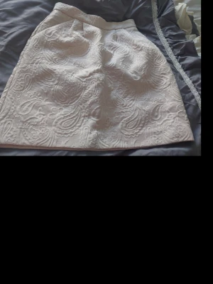 Vit kortkjol med broderat mönster - Säljer en vit kortkjol med snyggt broderat paisleymönster. Kjolen har en lätt A-linjeform och är tillverkad i ett strukturerat tyg som ger en exklusiv känsla. Perfekt för dig som vill ha något stilrent men ändå unikt.