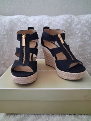 Svarta kilklackssandaler Michael Kors - Snygga svarta sandaler från Michael Kors med hög kilklack i flätad jute. Skorna har breda remmar, öppen tå och dragkedja i guld framtill. Perfekta för att lyfta din sommarstil med en trendig och bekväm look.