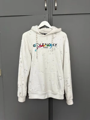 Givenchy hoodie med ”slitningar” - Säljer en vit hoodie från Givenchy med slitningar och broderad logga i flera färger på bröstet. Hoodien har huva och en stor magficka. Materialet är mjukt och skönt. Perfect för dig som älskar fashion! Storleken är XL med passar mer som M/L