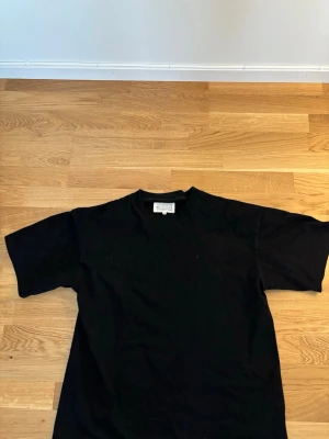 Svart t-shirt från Maison Margiela - En stilren svart t-shirt från Maison Margiela med klassisk rund halsringning och korta ärmar. T-shirten är i mjuk bomull och har en minimalistisk design utan tryck eller mönster. Perfekt för en clean och modern look.