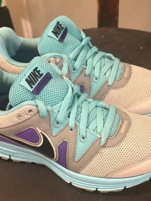 Nike sneakers i turkos och lila - Nike sneakers med mesh-ovandel i turkos och grått, detaljer i lila och svart Swoosh. Skorna har turkosa skosnören och en bekväm, vadderad insida. Snygg färgkombo och sportig design som sticker ut.