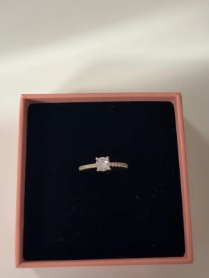 Guldfärgad ring med stor sten - Snygg guldfärgad ring med en stor, rund klar sten i mitten och små gnistrande stenar längs bandet. Perfekt för dig som gillar bling och vill sticka ut. Ringen levereras i en söt rosa ask.