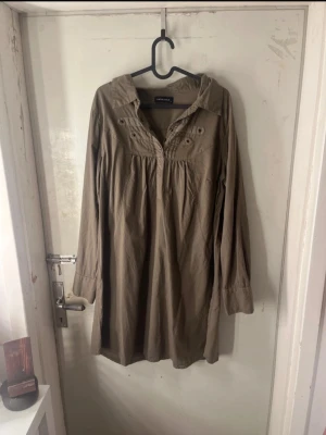 Klänning - Snygg brun klänning från Mamalicious med krage och dekorativa knappar vid bröstet. Blusen har lång ärm och är tillverkad i ett mjukt, vävt material som ger en avslappnad och luftig känsla. Perfekt för en stilren och bekväm look. Perfekt till sommar!