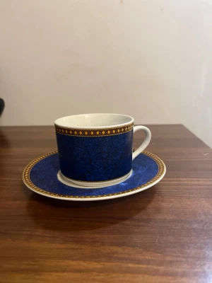 Blå kaffekopp med fat, porslin - Säljer en elegant blå kaffekopp med matchande fat i porslin. Koppen och fatet har en djupblå färg med dekorativ guldkant och mönster i gult och rött längs kanten. Perfekt för dig som gillar stilrena detaljer till fikastunden.