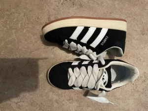 Svarta Campus sneakers med vita ränder - Svarta sneakers från Campus med klassiska vita ränder på sidorna och vit sula. Skorna har breda vita skosnören och en brun yttersula i gummi. Ovandelen är i textil och syntet, med låg profil och sportig look.