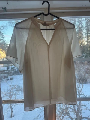 Blus - Snygg krämvit  blus i transparent material med v-ringning och korta ärmar. Blusen har lätt plisserade detaljer vid axlarna och en lös, luftig passform. Perfekt att styla lager på lager för en trendig look.