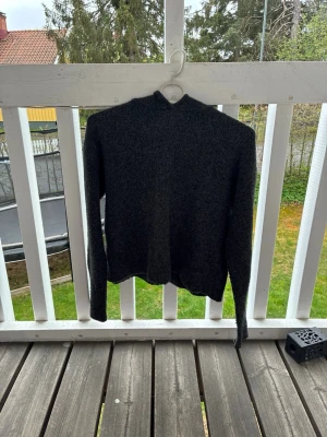 Mörkgrå stickad tröja med hög krage(czarno-szary sweter dzianinowy) - Mysig mörkgrå stickad tröja med hög krage och långa ärmar. Tröjan har en rak passform och är tillverkad i ett mjukt, ull-liknande material. Perfekt för kyliga dagar och enkel att matcha med jeans eller kjol.