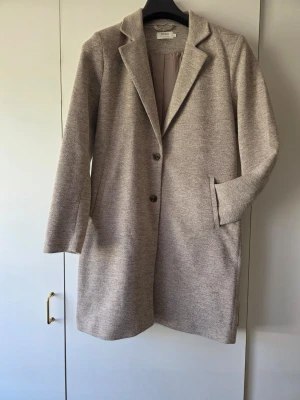 Beige kappa från Only med knappar - Stilren beige kappa från Only med två knappar framtill och klassisk krage.  Färgen är beige-grå eller grå-beige. Kappan har raka ärmar, två fickor och är fodrad på insidan. Perfekt för dig som gillar en enkel och tidlös look. Materialet är mjukt och har en lätt struktur.