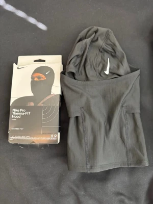 Nike Pro Therma-FIT svart huva - Nike Pro Therma-FIT Hood i svart, tillverkad i ett stretchigt och värmande material. Huvan har en tight passform och är designad för att hålla dig varm under träning utomhus. Swoosh-logga framtill och diskret etikett bak.