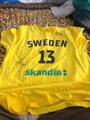 Gul Sverige matchtröja nr 13 - Gul matchtröja från Sverige med tryckta detaljer i blått, nummer 13 på ryggen och texten SWEDEN. Tröjan har två autografer och Skandia-logga framtill. Tillverkad i lätt och bekvämt material, perfekt för sport eller samling.