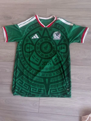 Mexiko landslagströja Adidas grön - Säljer en grön Mexiko landslagströja från Adidas med aztekiskt mönster och broderad landslagsemblem. Tröjan har vita och röda detaljer vid krage och ärmslut samt klassiska Adidas-ränder på axlarna. Tillverkad i lätt och ventilerande material, perfekt för fotboll.