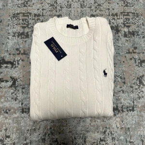 Vit kabelstickad tröja  - Vit kabelstickad tröja från med rund halsringning och broderad marinblå logga på bröstet. Tröjan har långa ärmar och ribbade muddar vid ärmslut och nederkant. Perfekt för en stilren look.