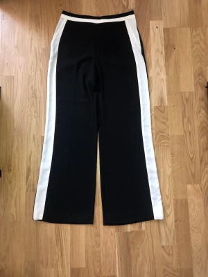 Svarta kostymbyxor med vita revärer Nya  h&m - Svarta kostymbyxor med raka ben och breda vita revärer längs sidorna och midjan för kontrast. Byxorna har medelhög midja och en stilren look. Materialet är mjukt och lätt, perfekt för dig som gillar en modern men unik stil. Ny med prislapp borta