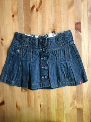 Mörkblå jeanskjol med knappar Esprit  - Snygg mörkblå jeanskjol från Esprit med knappar framtill och veckade detaljer. Kjolen har bred linning, bälteshällor och en liten broderad logga. Perfekt för dig som gillar en klassisk men trendig look.