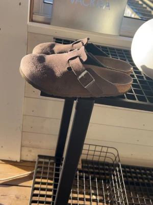 Birkenstock Boston - Säljer ett par bruna Birkenstock Boston tofflor i mocka med justerbart spänne i metall. Klassisk slip-in modell med rundad tå och bekväm sula. Perfekta för en avslappnad och trendig stil.