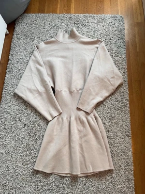 Beige stickad klänning från Zara - Snygg beige stickad klänning från Zara med hög krage och långa ärmar. Klänningen har en markerad midja med ribbad detalj och ett mjukt, stretchigt material som ger en skön passform. Perfekt för dig som gillar stilrena och bekväma plagg.