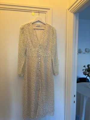 Blommig långklänning från Adoore - Superfin långklänning från Adoore i ljusgul ton med små blommor. Klänningen har v-ringning, långa ärmar och knappar framtill. Perfekt för sommarens alla tillfällen och har en feminin, romantisk stil.