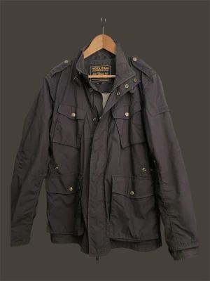 Woolrich Field jacket - Stilren mörkblå Field jacket från Woolrich med fyra stora fickor framtill, axelklaffar och dragkedja samt knappar. Jackan har en klassisk krage och är tillverkad i ett tåligt material som ger en cool och tidlös look.