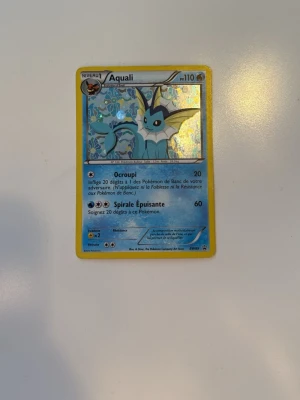 Pokémonkort Aqualis Vaporeon holo - Säljer ett Pokémonkort med Aqualis (Vaporeon) motiv. Kortet har en glittrig holografisk yta och är tryckt på klassiskt Pokémon-kartongmaterial. Framsidan är blå och gul med fransk text och baksidan har den ikoniska Pokéball-designen.