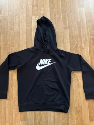 Svart Nike hoodie med logga - Svart hoodie från Nike med stor vit logga på bröstet. Klassisk modell med huva och känguruficka framtill. Tillverkad i mjukt material som är skönt att ha på sig. Perfekt för en sportig och avslappnad stil.
