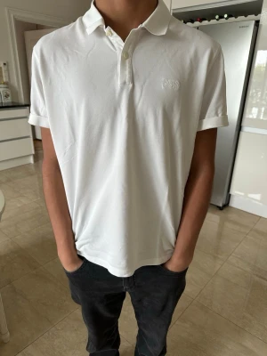 Vit piké t-shirt från Boss - Vit piké t-shirt från Boss med klassisk krage och knappar framtill. T-shirten har korta ärmar och är gjord i bomull. Diskret broderad logga på bröstet för en clean och stilren look.