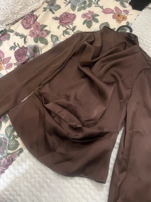 Brun satinblus  - Snygg brun blus i satin med lång ärm. Blusen har en elegant, figurnära passform och hög krage. Perfekt för dig som gillar stilrena och trendiga plagg med en lyxig känsla.