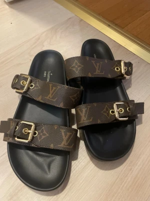 sandaler med monogram - Snygga sandaler med klassiskt brunt monogrammönster och två justerbara remmar med guldfärgade spännen. Svart fotbädd i mjukt material för extra komfort. Perfekta för en trendig och lyxig look.