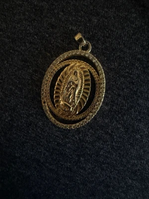 Guldigt halsband med religiös medaljong - Säljer ett ovalt halsbandshänge i guldfärg med en detaljerad religiös motiv i mitten, omgiven av en glittrande ram. Hänget har en klassisk look och passar perfekt till en kedja. Materialet ser ut att vara metall med stenar runt kanten för extra bling.