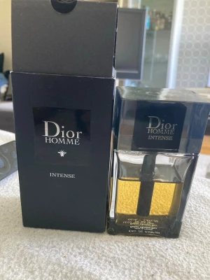 Dior Homme Intense Eau de Parfum - Dior Homme Intense Eau de Parfum är en lyxig herrdoft i en stilren, fyrkantig glasflaska med svart lock. Parfymen har en gyllene färg och kommer i en elegant svart kartong. Flaskan rymmer 100 ml och utstrålar exklusivitet och modern design.