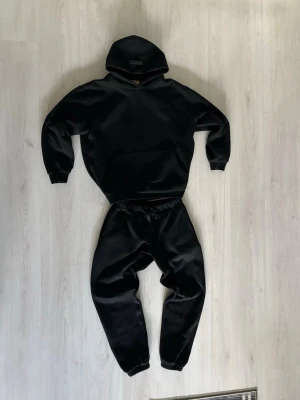 Hoodie & byxor i märket ESSENTIALS - Säljer ett ser ESSENTIALS med resår i midjan och vid bensluten. settet har en avslappnad passform och är tillverkade i ett mjukt material som känns skönt mot huden. Perfekta för chill dagar eller när du vill vara bekväm.