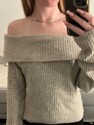 Beige off shoulder stickad tröja - Trendig beige stickad tröja med ribbad struktur och off shoulder-modell. Långa ärmar och bred krage som vilar över axlarna. Perfekt för en stilren och avslappnad look.