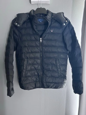 Svart jacka från Gant med huva - Snygg svart pufferjacka från Gant med avtagbar huva och dragkedja framtill. Jackan har två sidofickor och diskret logga på bröstet. Perfekt för kalla dagar och enkel att matcha med olika outfits. Tillverkad i lätt och värmande material. Storlek 164, 12-14 år 