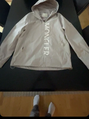 Grå vindjacka från Moncler med huva - Snygg grå vindjacka från Moncler med stor logga framtill. Jackan har huva, dragkedja, två fickor med dragkedja och justerbar nederkant. Perfekt för blåsiga dagar och enkel att matcha med olika outfits.