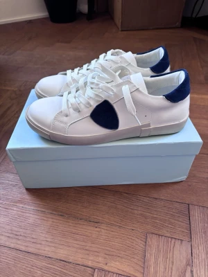 Philippe model sneakers - Snygga vita sneakers med blå detaljer i mocka på sidorna och hälen. Skorna har klassisk låg profil, vita snören och en stilren design som passar till det mesta. Ovandelen är i slätt läder och insidan är fodrad för extra komfort. Skor påse medföljer